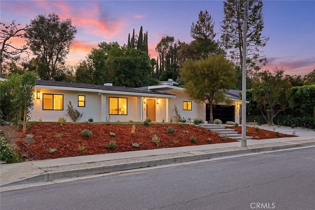 Twilight elevation of 4615 El Caballero Drive, a 1961 mid-century modern in Tarzana, with low-pitched eaves and warm interior light against a sunset sky.